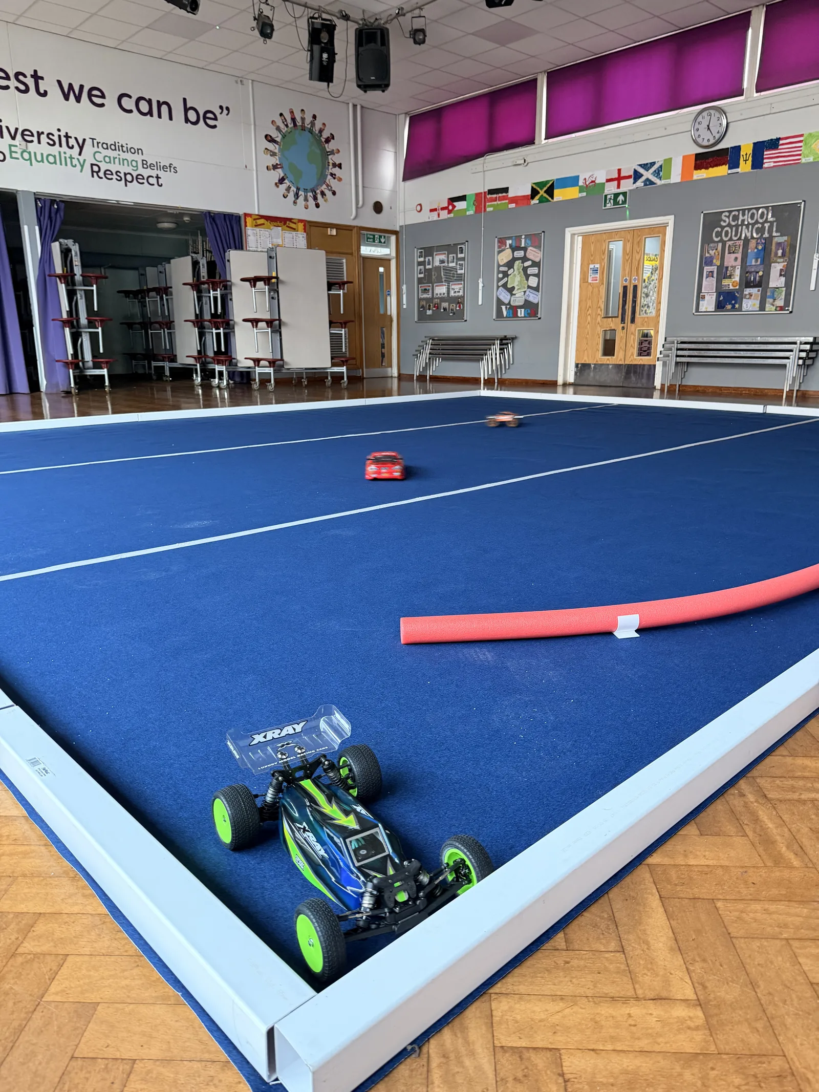 An RC buggy and a touring car on the blue track carpet inside the school hall, next to a white guttering edge and a pool noodle barrier